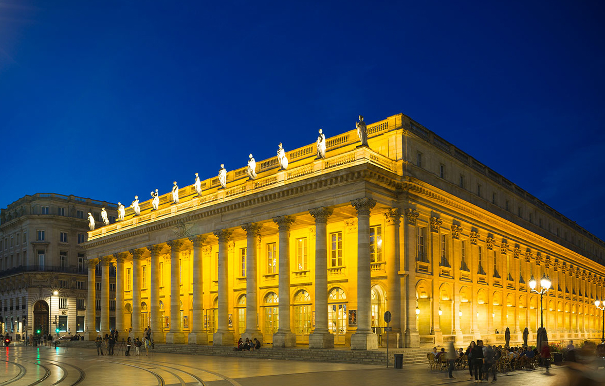 Prise de nuit la photo souligne l'architecture néo-classique du bâtiment mise en valeur par un jeu de lumières jaunes-orangées. On y voit la façade en pierre de théâtre avec ses douze colonnes corinthiennes soutenant une corniche avec les statues des douze muses. Les trois niveaux du bâtiments sont percés de fenêtres et de porte-fenêtre. Aux pieds des colonnes, quelques marches donnent sur une place zébrées de rails de tramways.