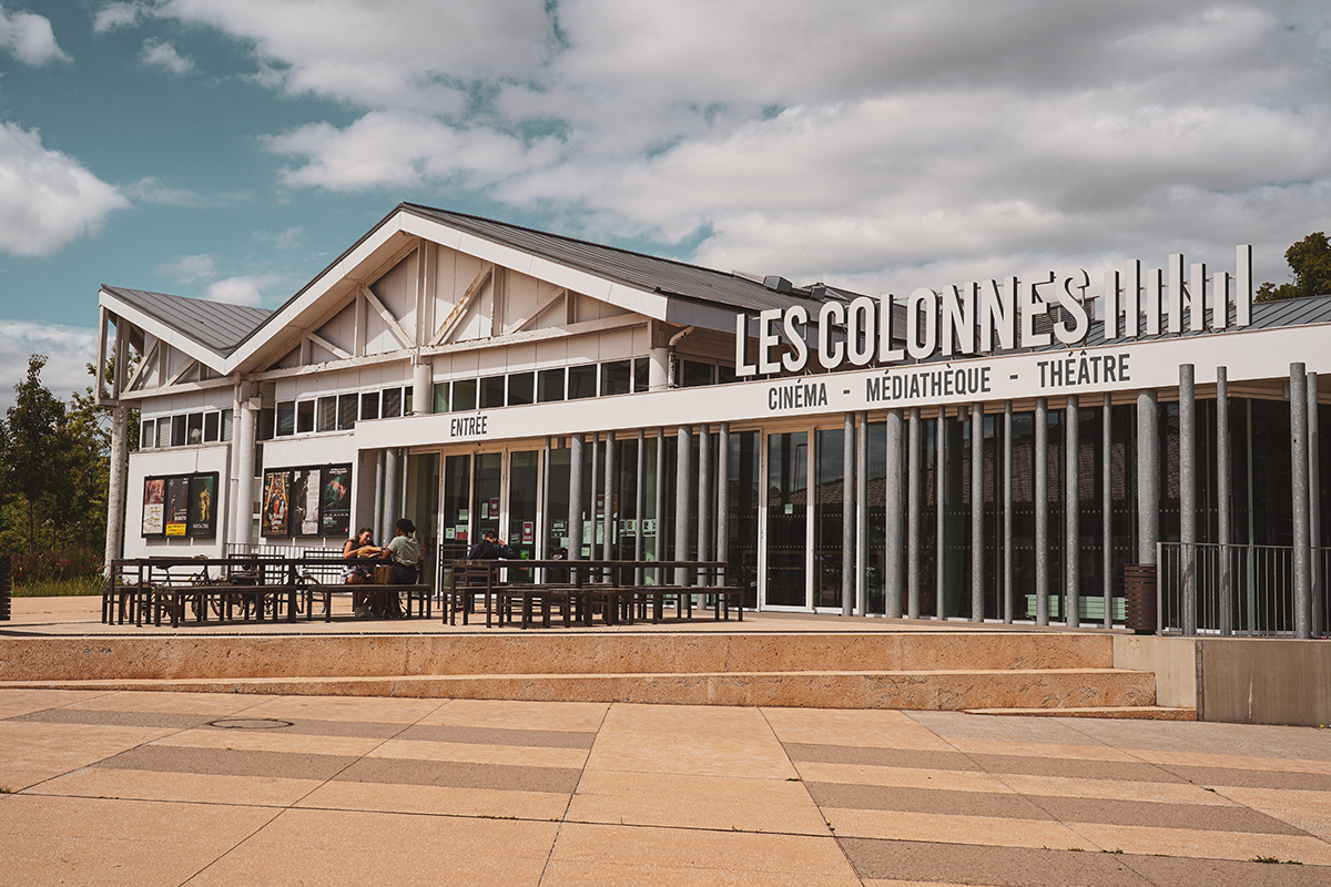 Bâtiment moderne en longueur avec un grand panneau indiquant "LES COLONNES CINÉMA - MÉDIATHÈQUE - THÉÂTRE". Le bâtiment a une façade blanche et de grandes fenêtres. Il y a des bancs et des tables en bois et en métal devant l'entrée principale.