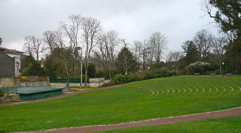 Grand espace herbeux en légère pente vers la gauche formant de légers espaliers. En bas de la pente, tout à fait à gauche, une scène verte surélevée à 1 mètre du sol. dans le fond des arbres.