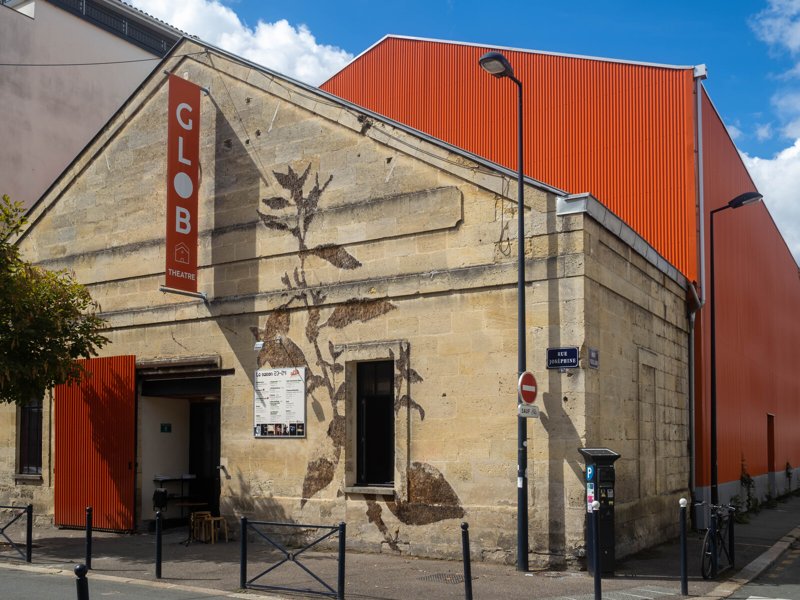 Bâtiment en pierres claires au toit pointu faisant le coin d'une rue. Au fronton pend une banderole verticale orange sur laquelle est écrit en blanc : "GLOB Théâtre". Un grande porte orange coulissante sert d'entrée. Sur la façade une fresque ocre représente une plante qui grimpe presque jusqu'au toit. Dans le prolongement de la bâtisse, un immense bâtiment peint également en orange. 